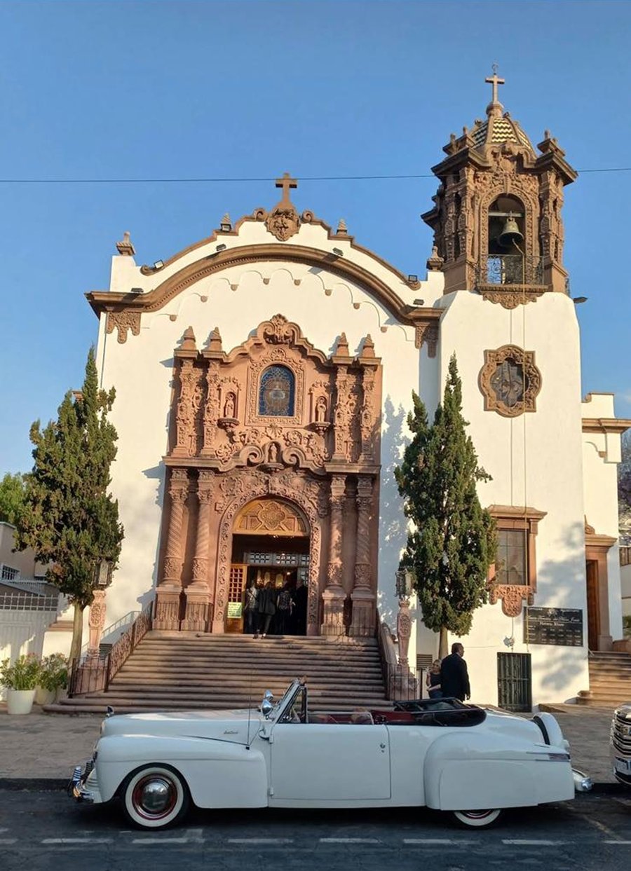 Fachada de la iglesia con un coche clásico blanco aparcado delante, al atardecer